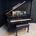Black grand piano with its lid open, a glossy finish, in a dark studio corner, with a cushioned bench in front.