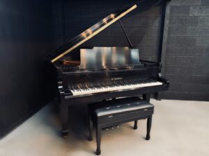 Black grand piano with its lid open, a glossy finish, in a dark studio corner, with a cushioned bench in front.