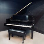 Black grand piano with a matching bench in a dark studio room; lid open revealing strings and frame. K. Kawai logo visible on the fallboard.