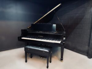 Black grand piano with a matching bench in a dark studio room; lid open revealing strings and frame. K. Kawai logo visible on the fallboard.