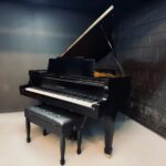 Black grand piano with open lid and matching bench in a dim rehearsal room.