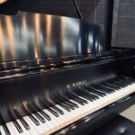 Shiny black grand piano with the keyboard visible, brand K. Kawai, set against a dark brick wall in a studio.