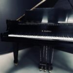 Black grand piano in a dark studio, showing its white keys and gold feet/pedals. The logo reads 'K. KAWAI' on the side of the piano.